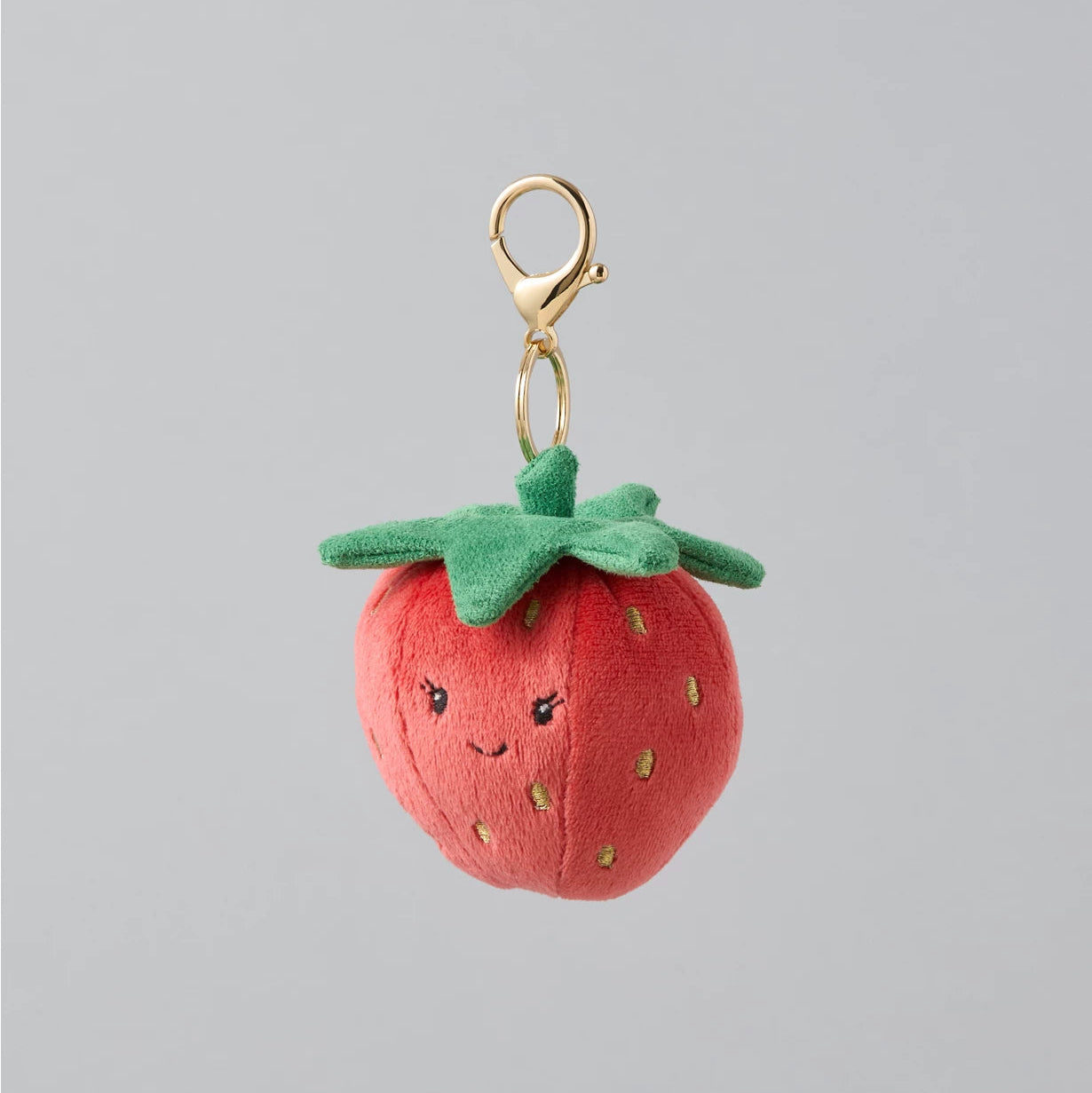 A small plush strawberry keychain charm with a gold clasp, featuring a cute embroidered face on a red body and green leaves, set against a solid gray background.