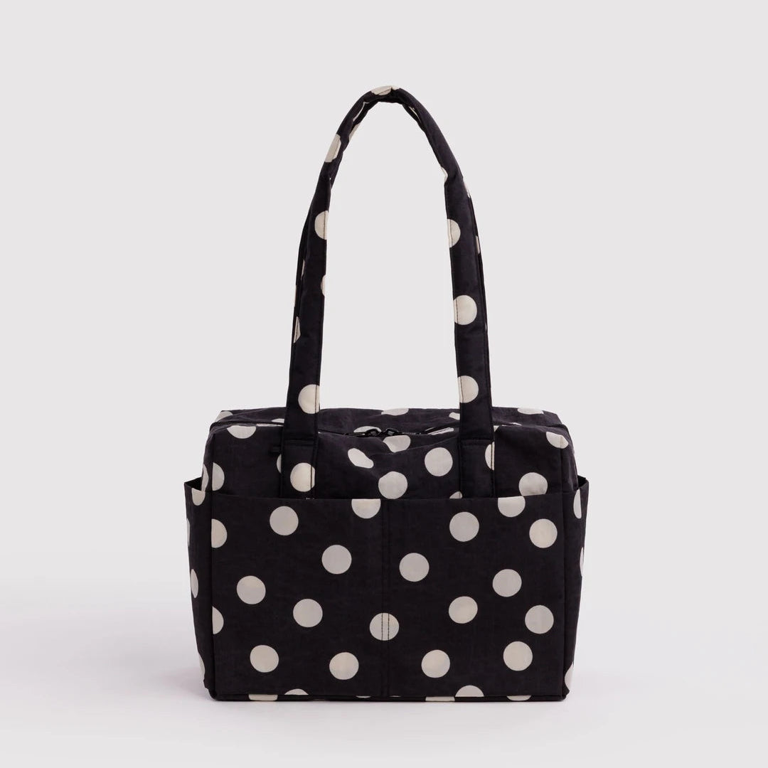 A black fabric tote bag with large white polka dots and dual top handles, photographed against a plain light gray studio background.